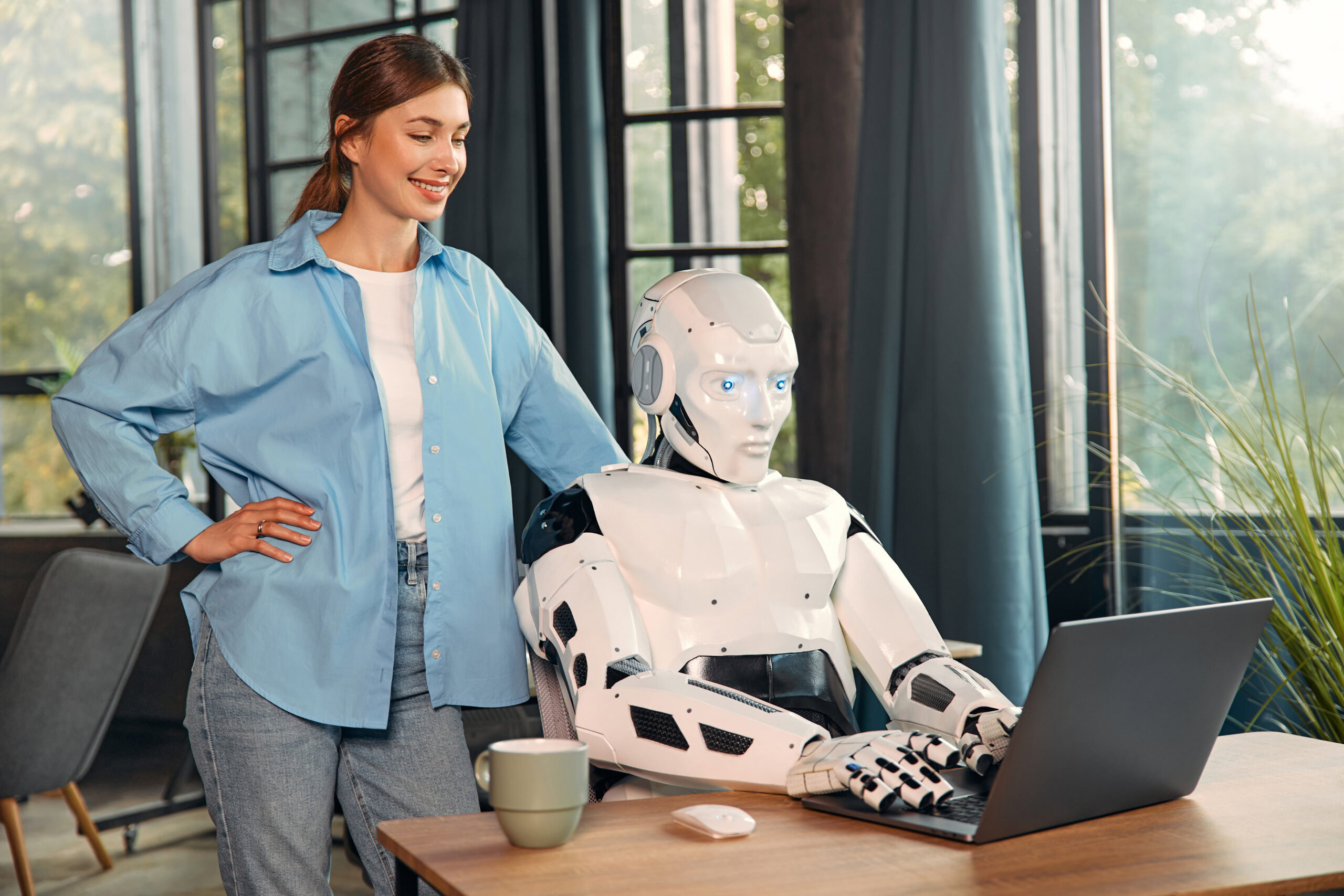 Young woman looking over a humanoid robot working on a laptop in a modern office. Collaboration between humans and Conversational AI.